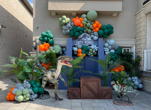 Jurassic dinosaur themed balloon backdrop in green, orange, and grey with life-size dinosaur props for birthday party