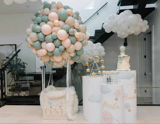 Whimsical hot air balloon-themed first birthday setup with pastel pink, sage, and cream balloon garland shaped like a balloon bouquet rising from a decorated gift box that says "ONE", with a white fluffy rug and custom cake display in the background.