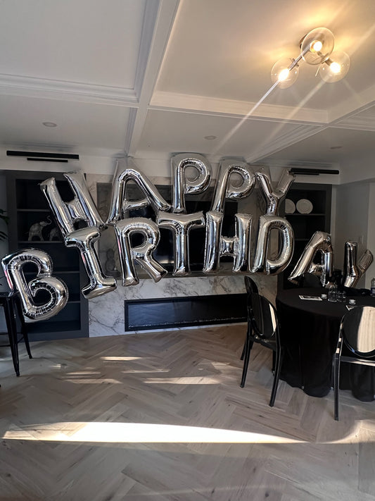 A grand silver foil 34-inch balloon letter arch spelling 'HAPPY BIRTHDAY' installed over a mantel for an elegant indoor event by WeBalloonz.