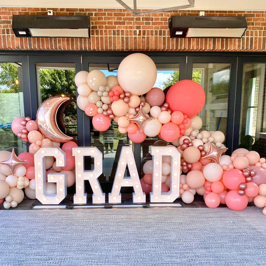 Graduation balloon garland in rose gold, nude, and peach tones with light-up marquee GRAD letters β custom outdoor decor setup for photo ops and grad parties.