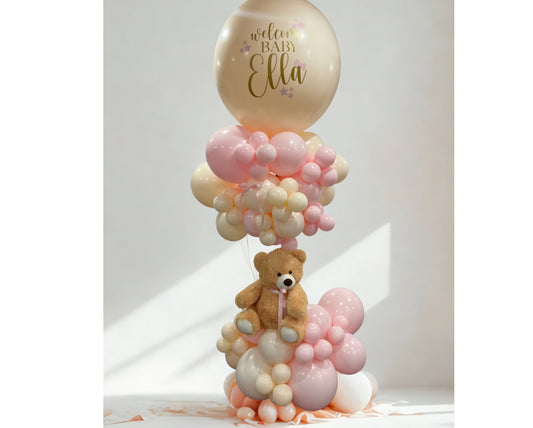 Balloon tower with teddy bear and personalized jumbo balloon that says “Welcome Baby Ella,” featuring soft pink, blush, and ivory balloons.