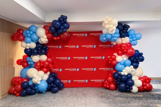 A festive balloon arch setup in front of a red backdrop printed with the SHOPPERS logo, featuring clusters of red, navy blue, and white balloons arranged on both sides of a central opening.