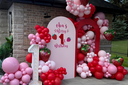 Custom organic balloon installation in shades of pink and red with strawberry foil balloons and a personalized arch backdrop for a girl's first birthday party in Toronto.