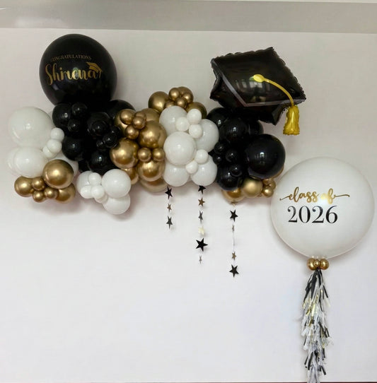 A 6ft organic graduation mini garland in black, white, and gold featuring a graduation hat balloon and a jumbo personalized helium balloon by WeBalloonz.