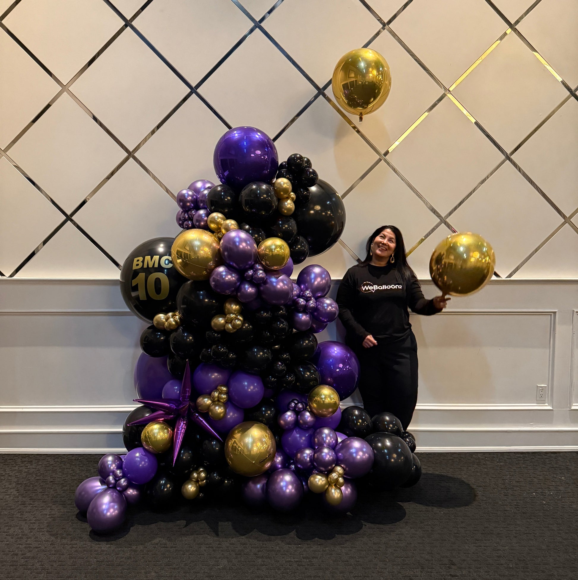 Stand alone balloon garland in black, purple, and gold for 10th anniversary at BMC event with WeBalloonz team member holding gold balloon