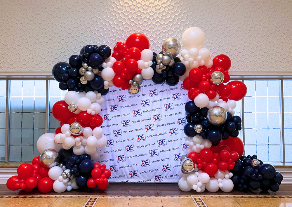 Full balloon garland in red, navy, white, and silver framing a branded step and repeat backdrop for corporate event