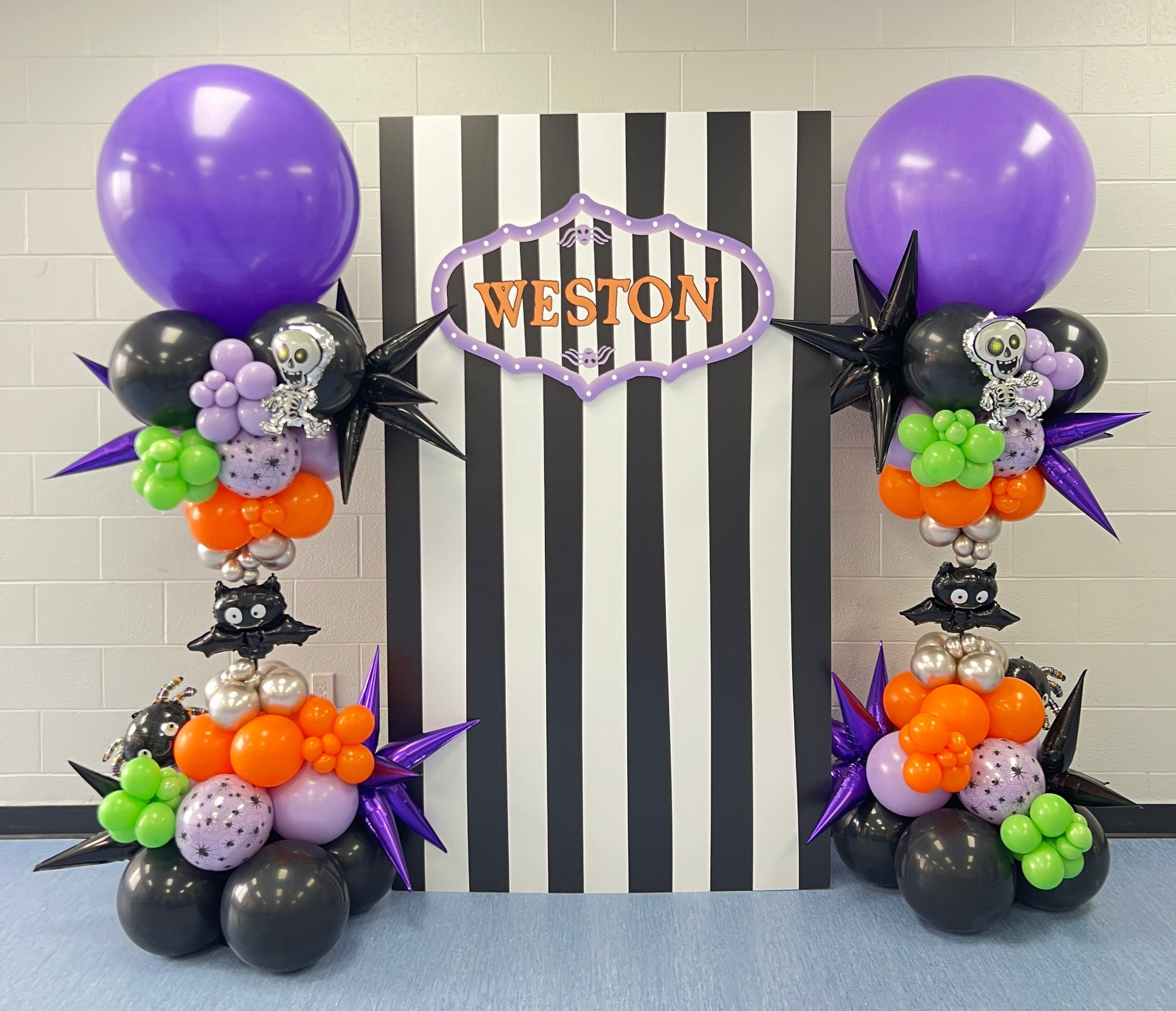 Halloween balloon towers with vibrant balloons in purple, orange, green, and black, flanking a black and white striped backdrop.