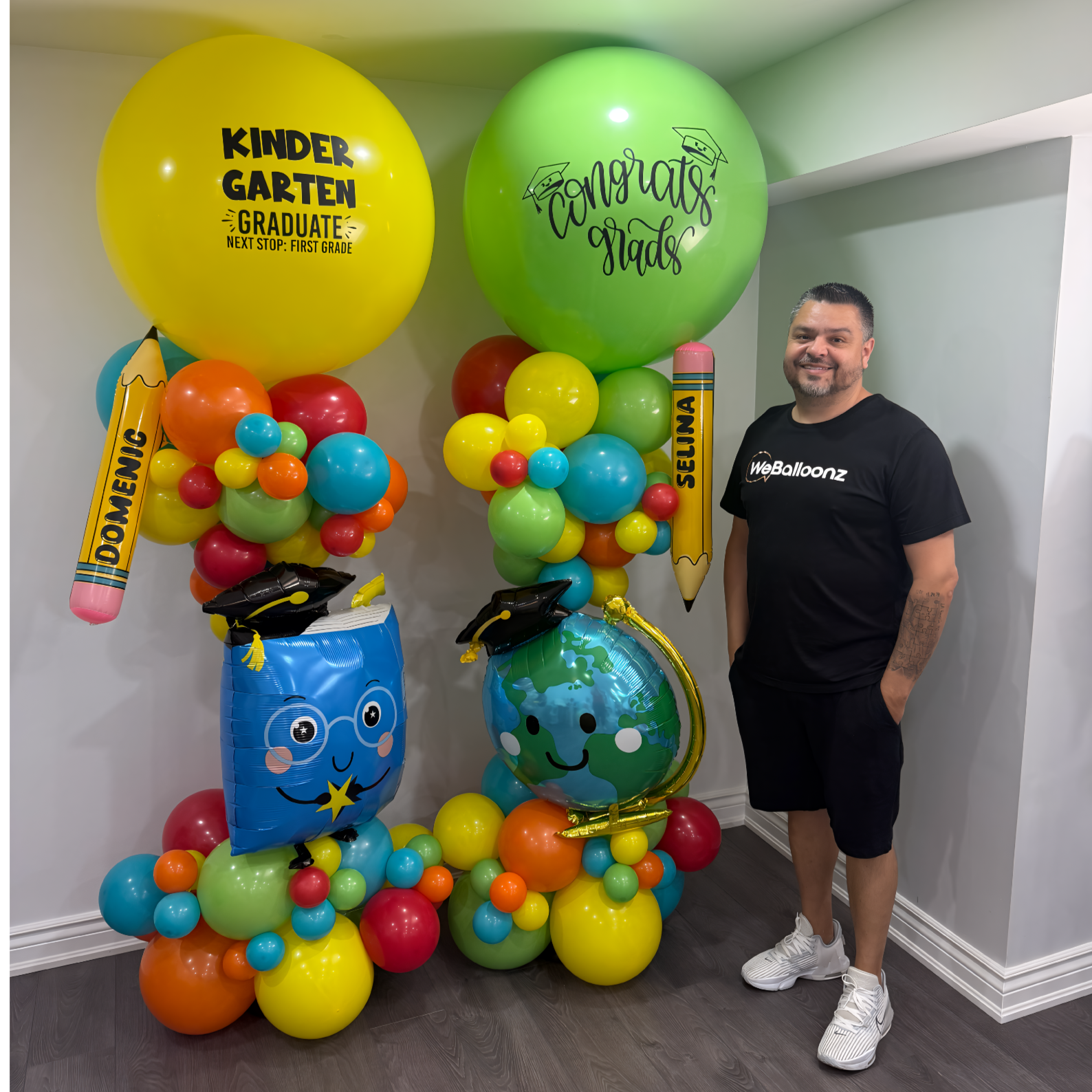 Man standing beside kindergarten graduation balloon towers with colorful balloons, jumbo personalized balloons, pencil foils, and graduation-themed characters for size reference