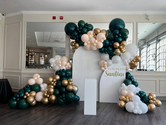 Emerald green, sand, and gold organic balloon garland with custom "God Bless Santino" arched backdrops for a luxury communion celebration by WeBalloonz