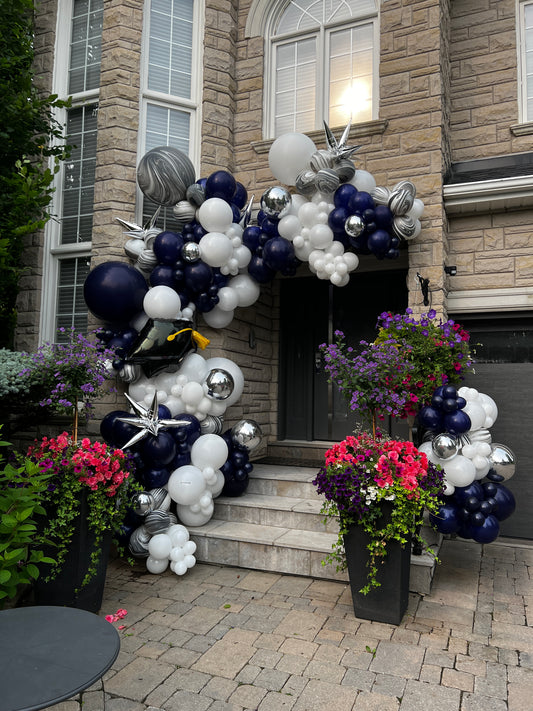 A grand 18ft luxury graduation entrance garland in navy, white, and silver chrome, featuring a jumbo graduation cap balloon and silver starburst accents framing a residential stone entrance in Vaughan by WeBalloonz
