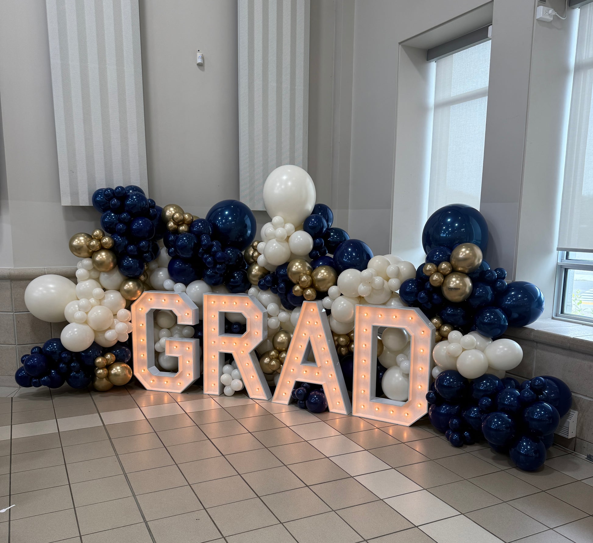 Large-scale indoor graduation backdrop featuring 3ft illuminated GRAD marquee letters and a bespoke navy, gold, and white organic balloon installation in Vaughan by WeBalloonz.