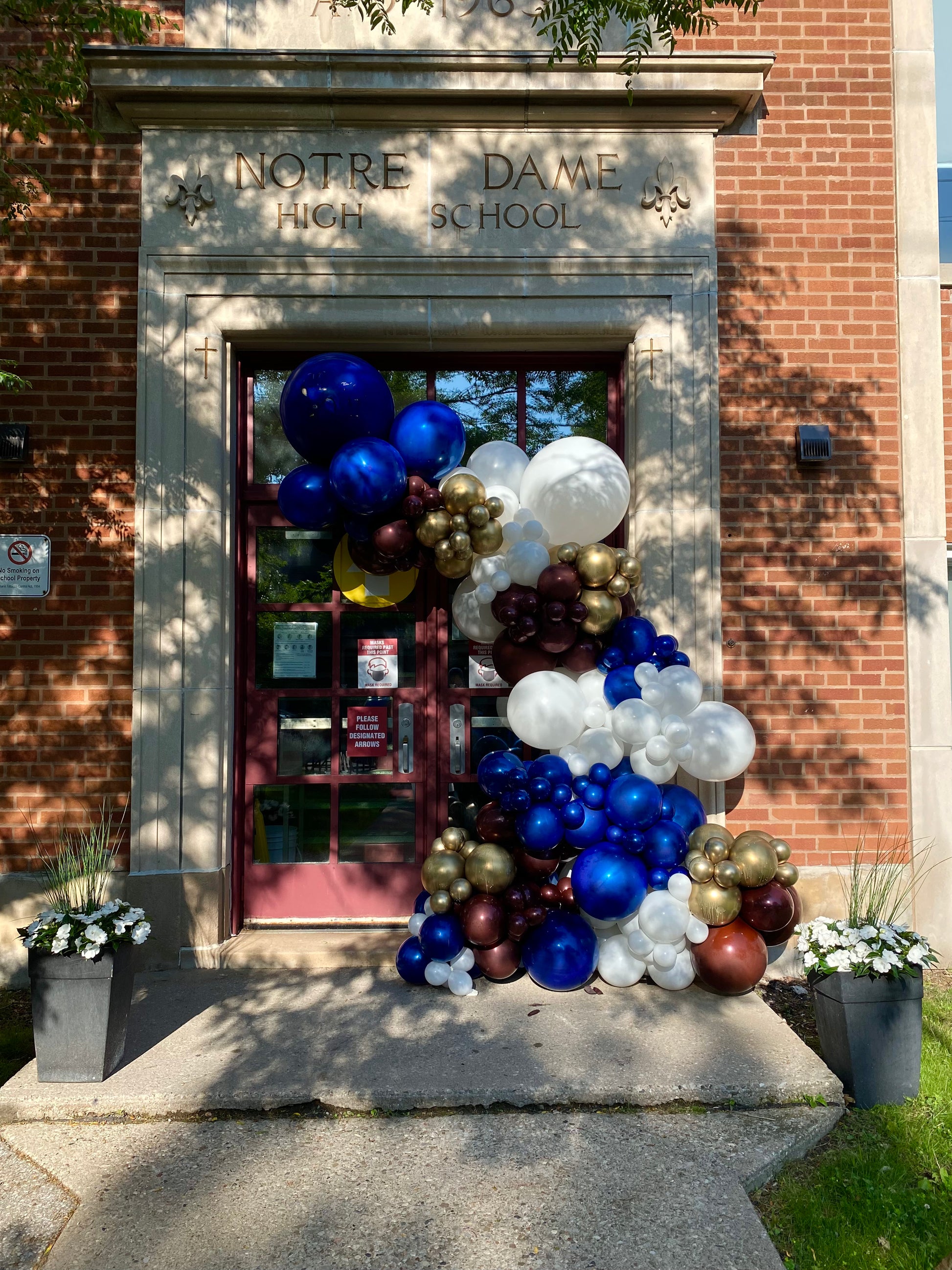 A massive high-density organic balloon garland in navy, gold, and white framing the main stone entrance of Notre Dame High School for a graduation celebration by WeBalloonz
