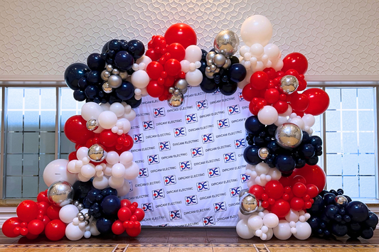 Custom red, white, and navy organic balloon arch framing a corporate step and repeat backdrop installation by WeBalloonz.