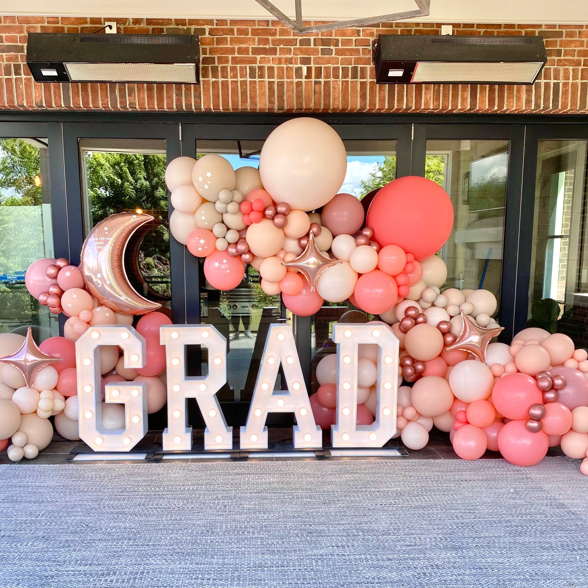 Graduation balloon garland in rose gold, nude, and peach tones with light-up marquee GRAD letters — custom outdoor decor setup for photo ops and grad parties.